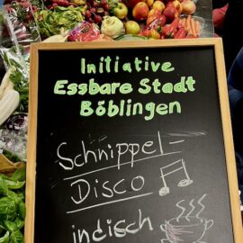 Die geretteten bunten Lebensmittel sind auf einem großen Tisch ausgebreitet und werden begutachtet. Im Vordergrund das Schild der Schnippeldisco: indisch Kochen - NAMASTE